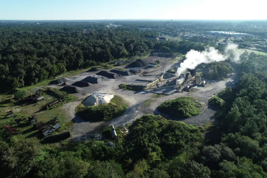Drone image of an active asphalt facility with visible stockpiles, smoke-emitting machinery, and surrounding forested areas supporting industrial operations