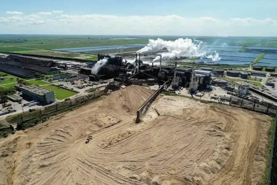 Aerial view of an sugar plant adjacent to a wooded area and pond, with visible stockpiles and facility infrastructure