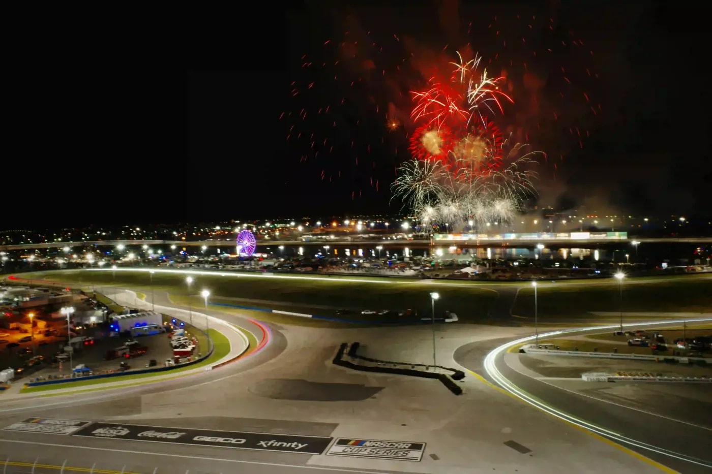 Nighttime shot of a brightly lit NASCAR racetrack with fireworks in the sky and a lit-up Ferris wheel in the distance