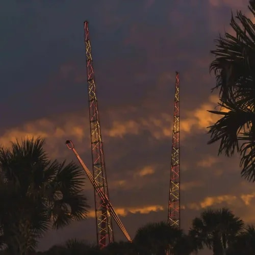 Amusement ride towers lit in red and yellow lights silhouetted against a dramatic sunset sky with palm trees