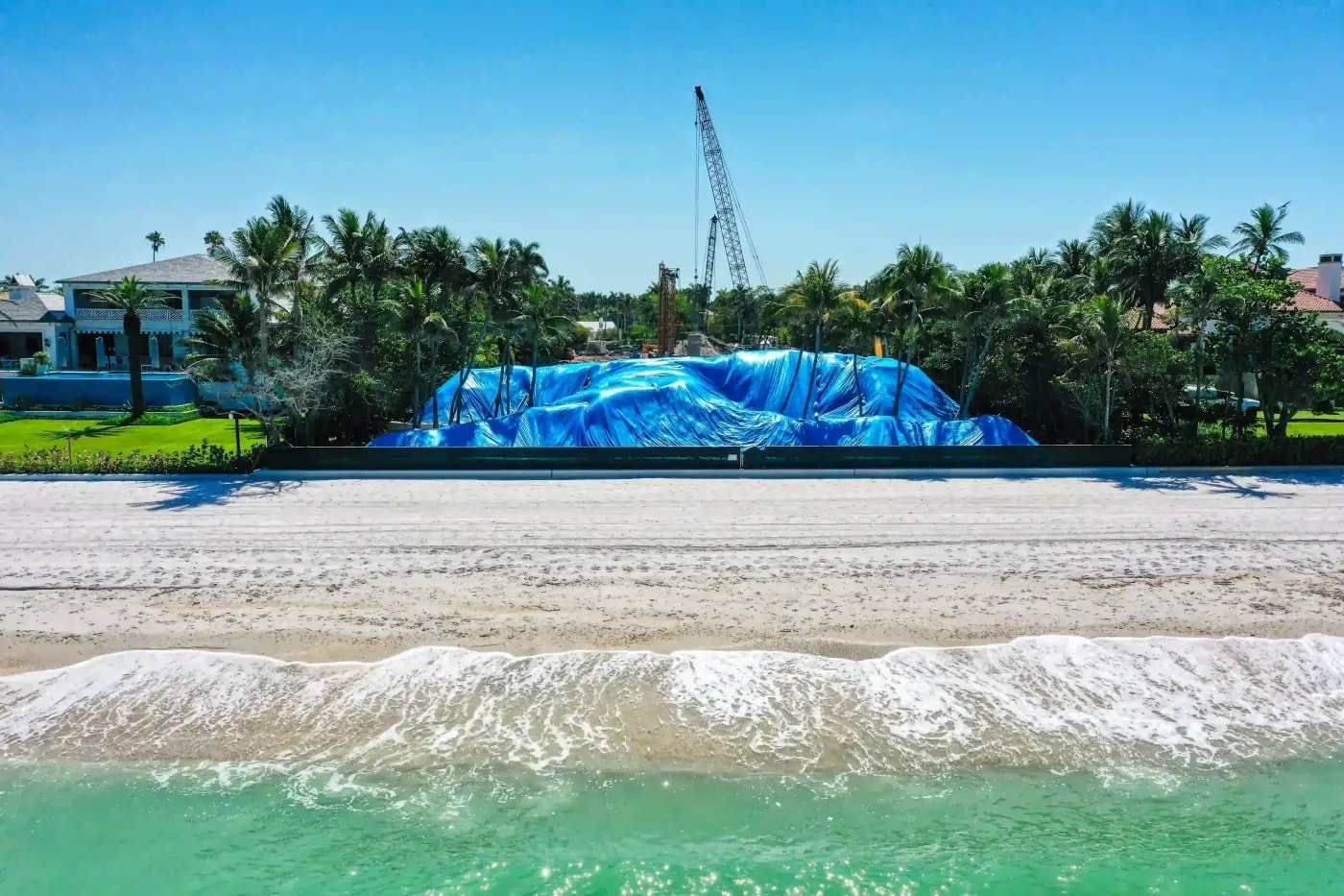Construction site with tarped materials and heavy equipment staged behind beachfront property near palm groves