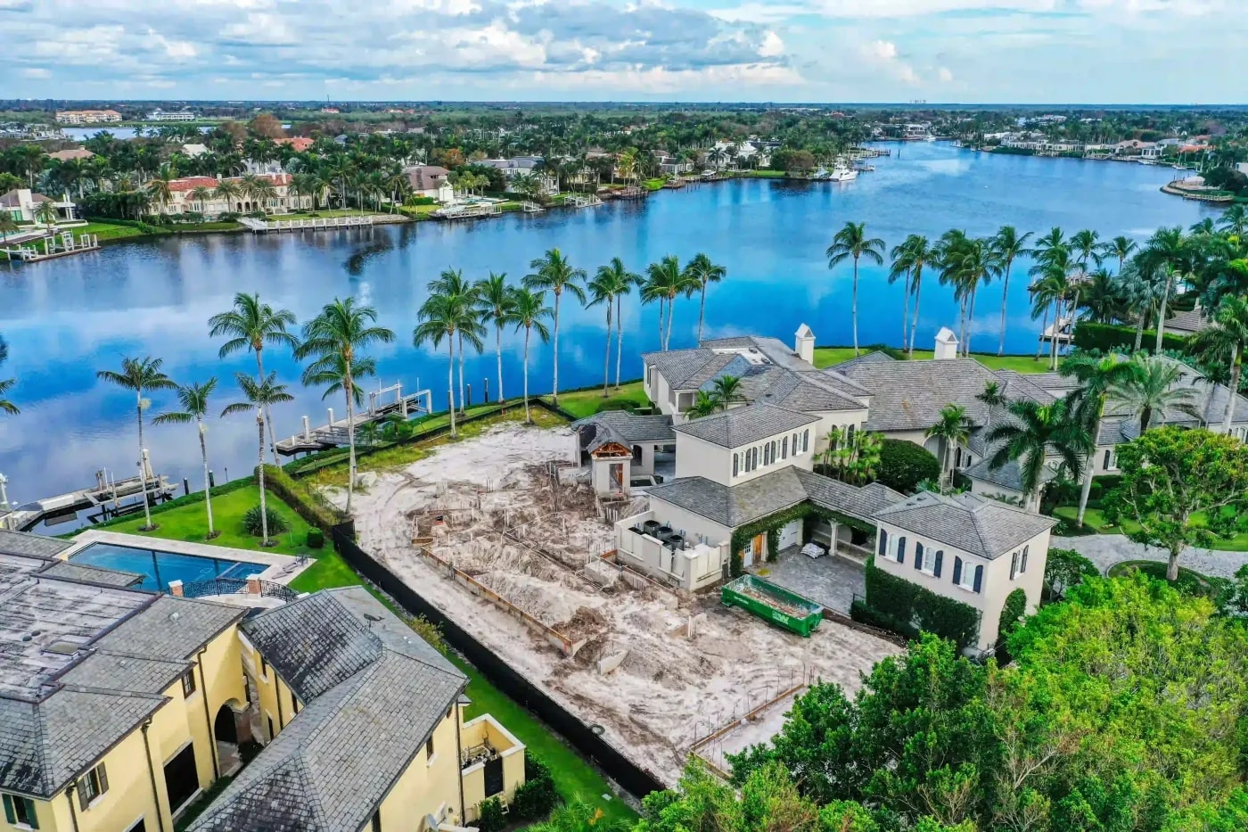 Drone view of foundation work for a luxury home near a canal, surrounded by palm trees and residential lots