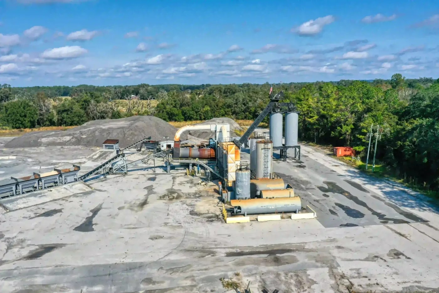 Ground-level drone shot of asphalt processing facility with material piles, silos, and conveyor systems in a cleared area