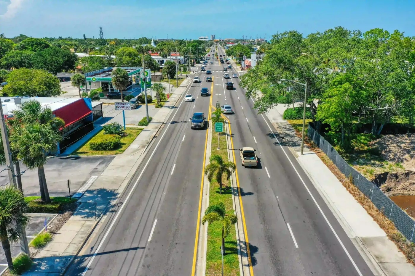 Street-level drone image of a multilane road with palm-lined median and moderate vehicle flow on a sunny day