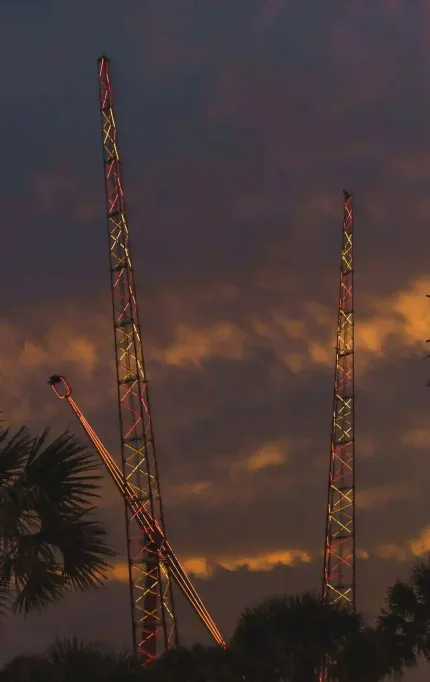 Amusement ride towers lit in red and yellow lights silhouetted against a dramatic sunset sky with palm trees