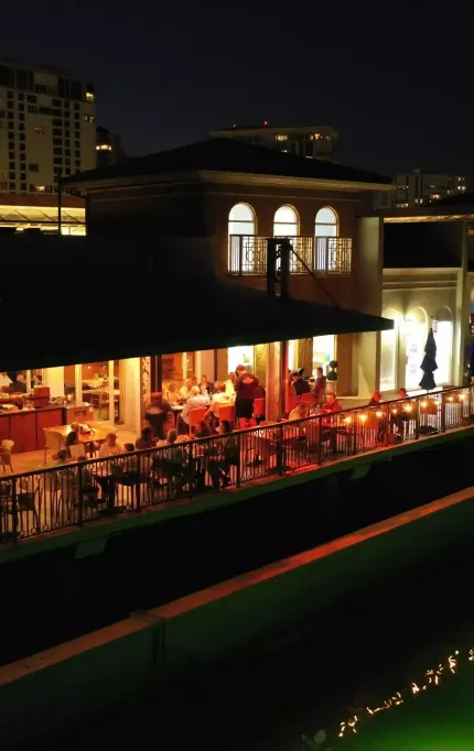 Evening drone view of open-air dining terrace beside a canal, with lights reflecting on the water and buildings in the background